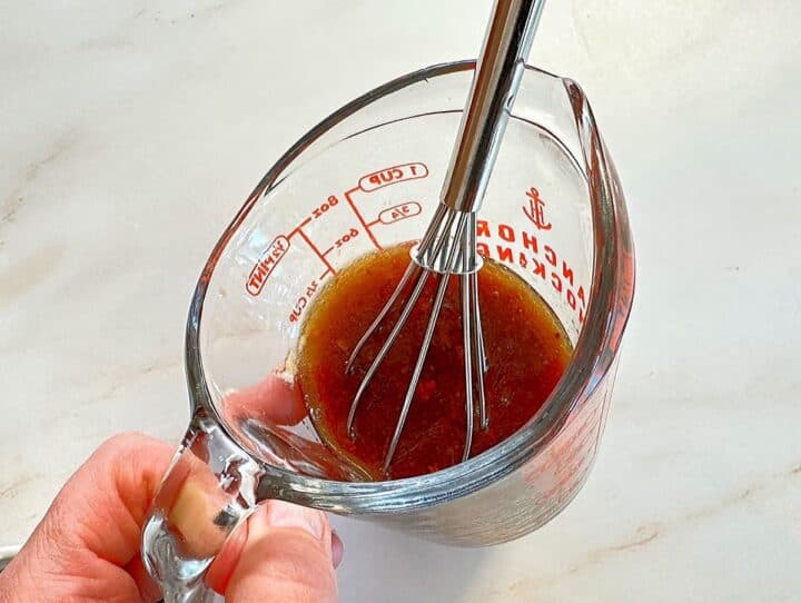 Brown soy marinade in clear glass measuring cup with whisk on cream marble countertop