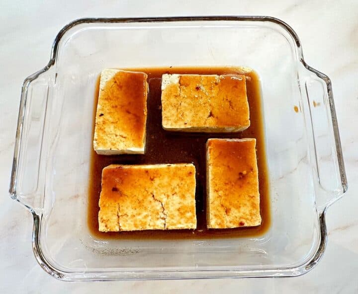 Four large pieces of tofu in square glass baking dish covered by brown marinade.