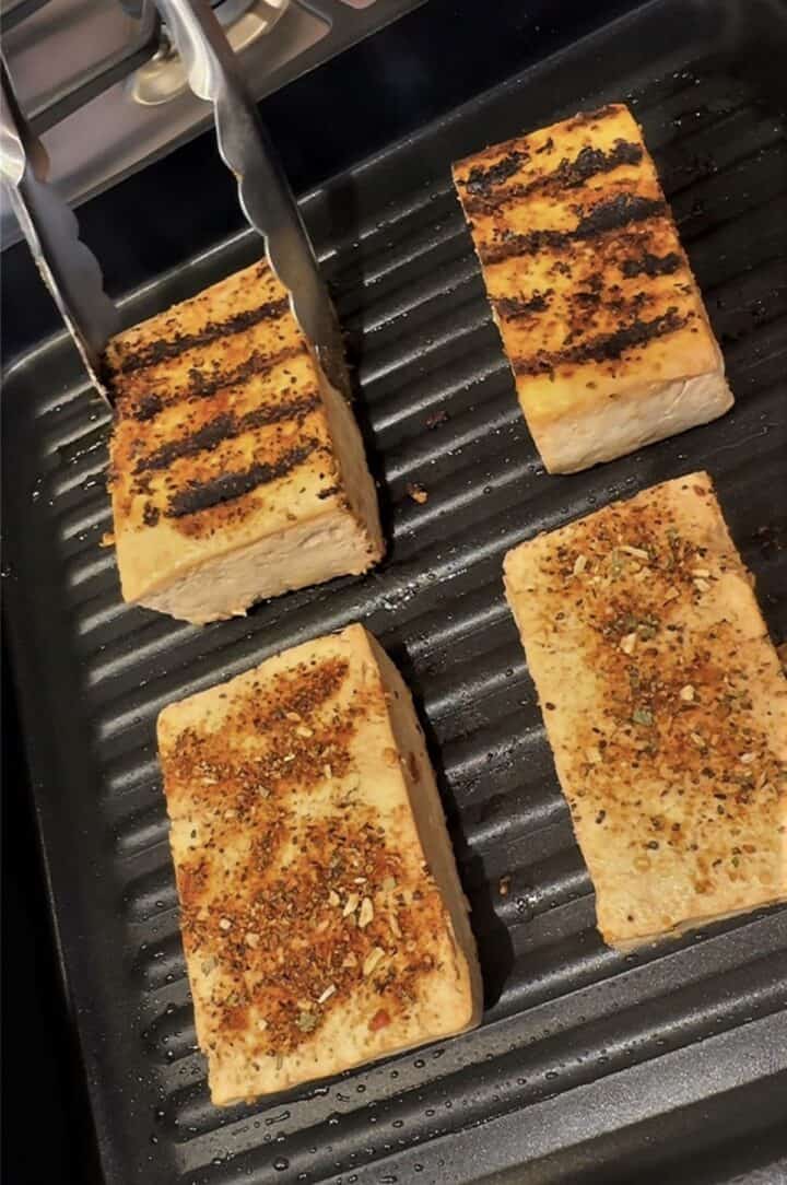 Tongs turning tofu steaks on a dark grill pan so you can see the grill stripes.