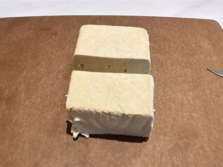 Off-white brick of tofu cut in half on brown cutting board.