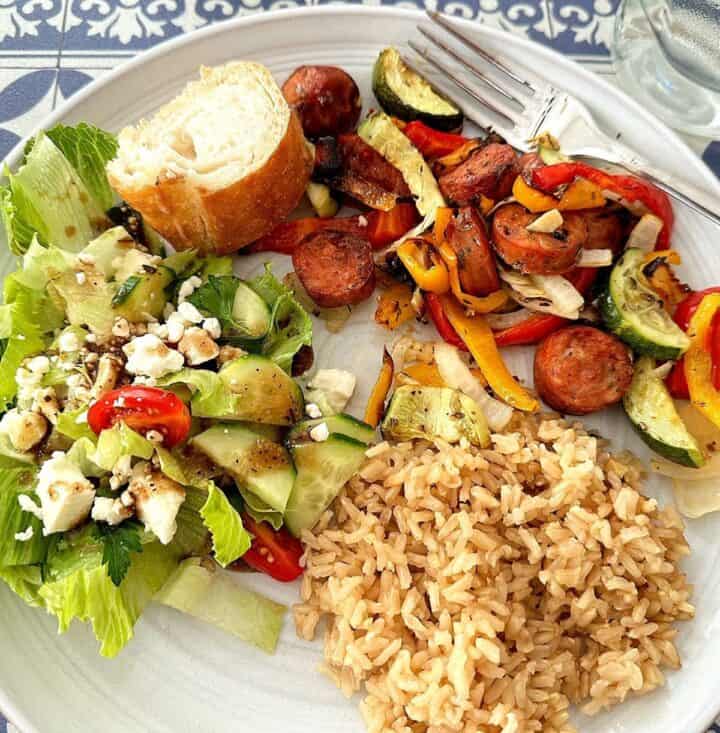 Round white plate with sausage coins, colorful peppers, zucchini, brown rice, green salad, and a slice of baguette