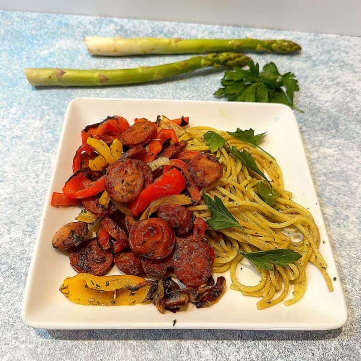 Square white plate with sausage coins, red and yellow peppers next to a pile of pasta on a bluish background