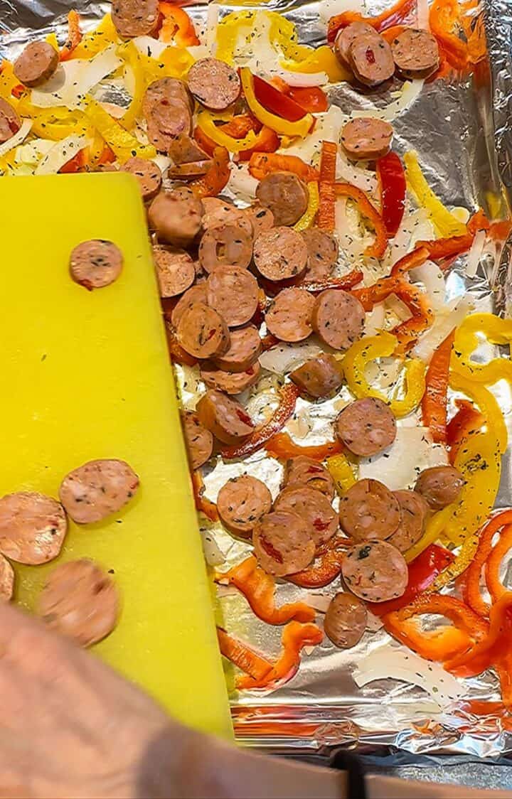 Adding sliced rounds of sausage to a sheet pan that contains strips of yellow and red peppers and sliced onion