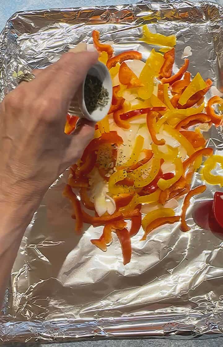 Round white ramekin of spices being sprinkled onto sliced red and yellow bell peppers and onions 