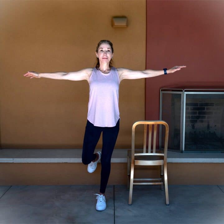 Woman in pink tank top and navy tights standing on one leg with a copper colored chair nearby.