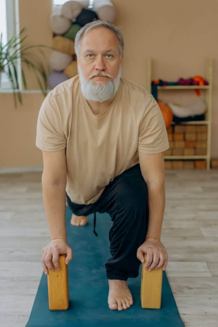 Man with white hair and beard wearing comfortable clothes on exercise mat in a lunge position with blocks