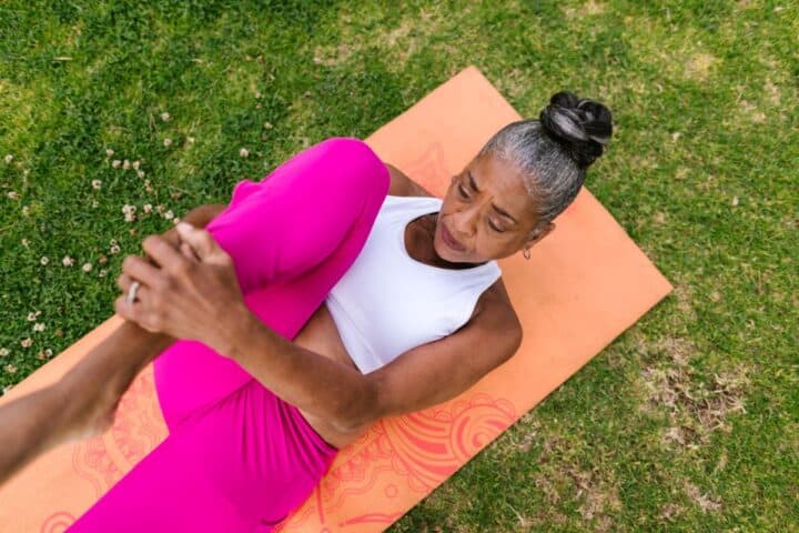 Older black woman wearing pink leggings and a white top lying on a salmon-colored mat hugging  knee to chest