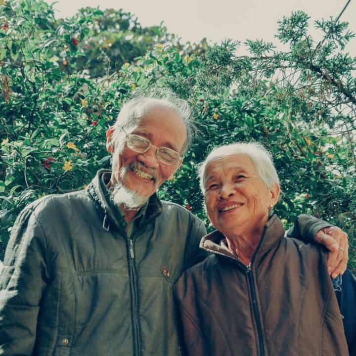 Older Asian couple (man and woman) standing close together and smiling amongst trees.