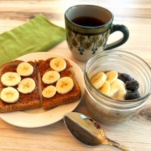 Breakfast scene on woodgrain with chia pudding topped with banana slices and blueberries, plus a slice of toast with sliced bananas and black coffee in a decorative mug.