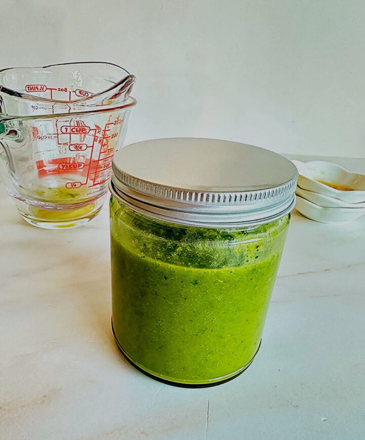 Round jar three-quarters full of green dressing on a light marble countertop with clear liquid measuring cups and white pinch bowls stacked in the background/