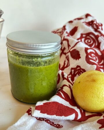 Clear round jar full of medium-green dressing next to a kitchen towel with a red rose pattern and a roundish lemon.