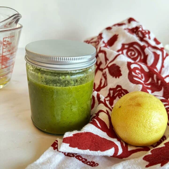 Clear round jar full of medium-green dressing next to a kitchen towel with a red rose pattern and a roundish lemon.