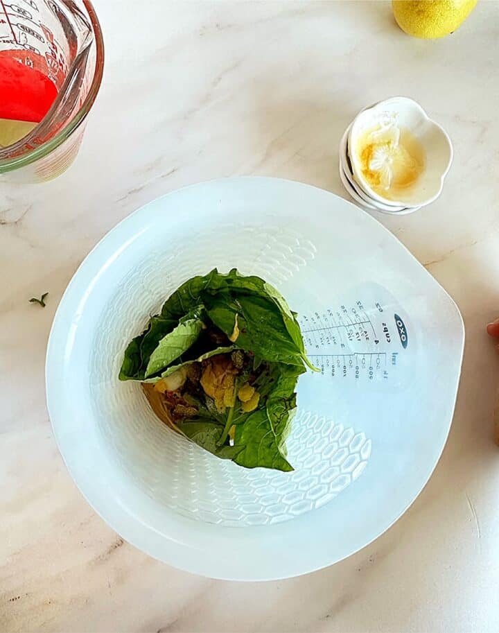 Bright green basil leaves, mustard, garlic clove, and other vinaigrette ingredients in an opaque oxo container with pinch bowls & measuring cups in the background.