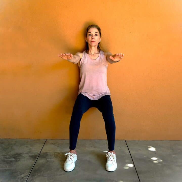 Woman wearing pink tank top and navy tights performing a wall sit against a golden stucco wall, reaching out with hands for balance