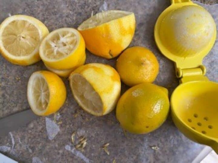 Whole lemons, plis lemons cut in half with a yellow citrus press alongside on a granite countertop.