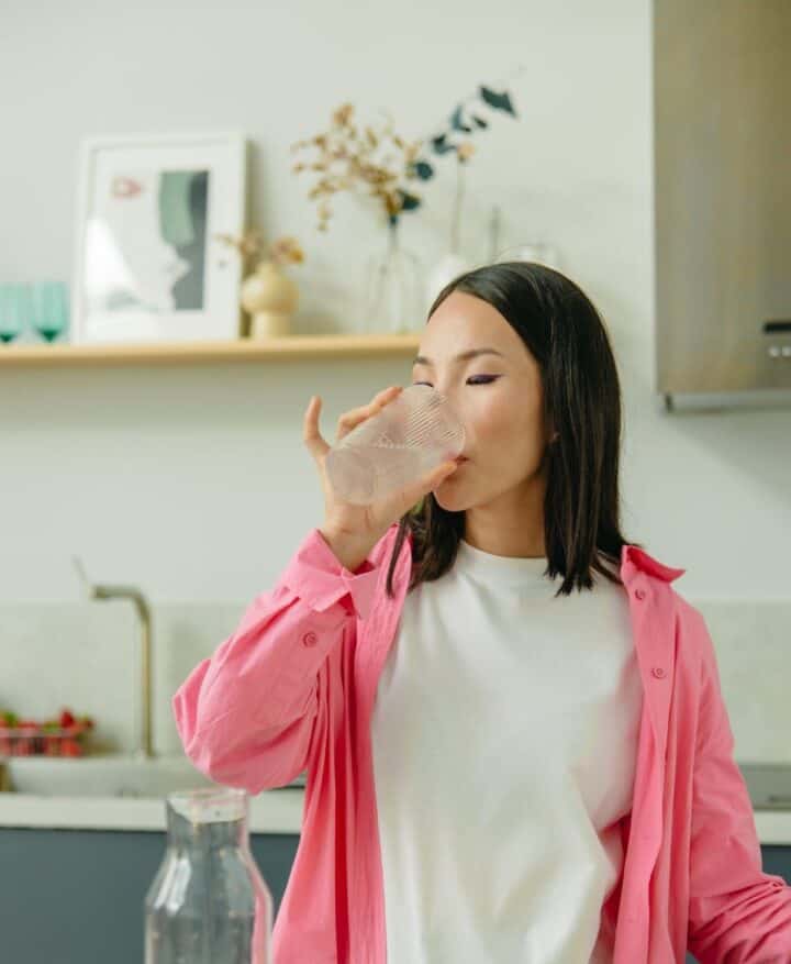 Asian woman in white shirt and pink jacket drinking a glas of water in a kitchen.