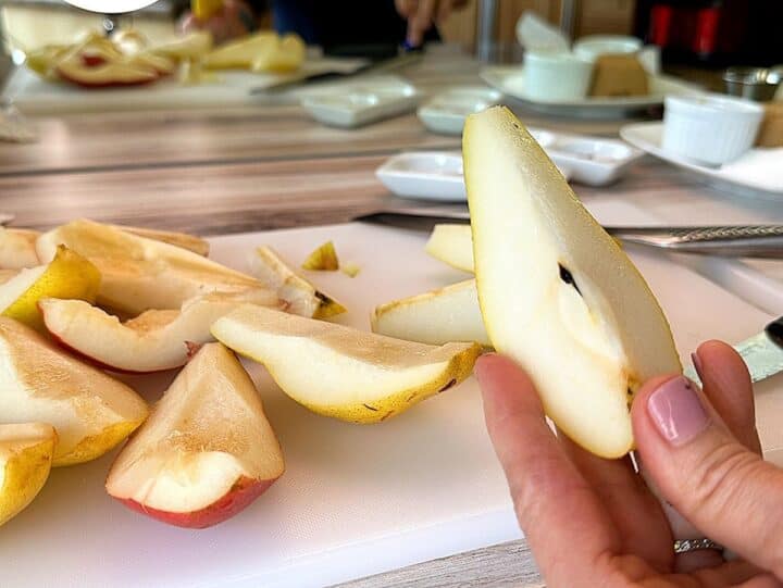 Pear quarter with core held in the foreground and white cutting board holding pear quarters with core removed in the background.