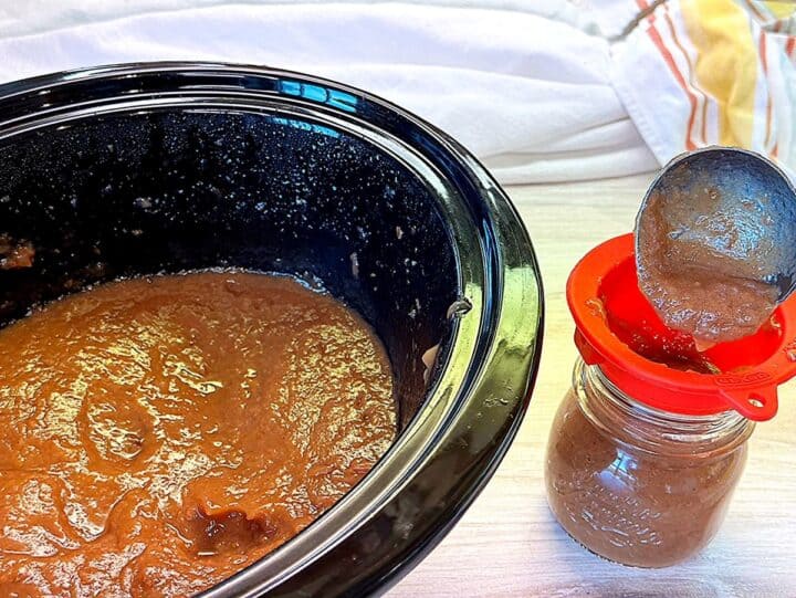 Caramel-colored pear butter in a black slow-cooker liner next to a jar that is being filled (by a ladle) with butter through a red funnel.