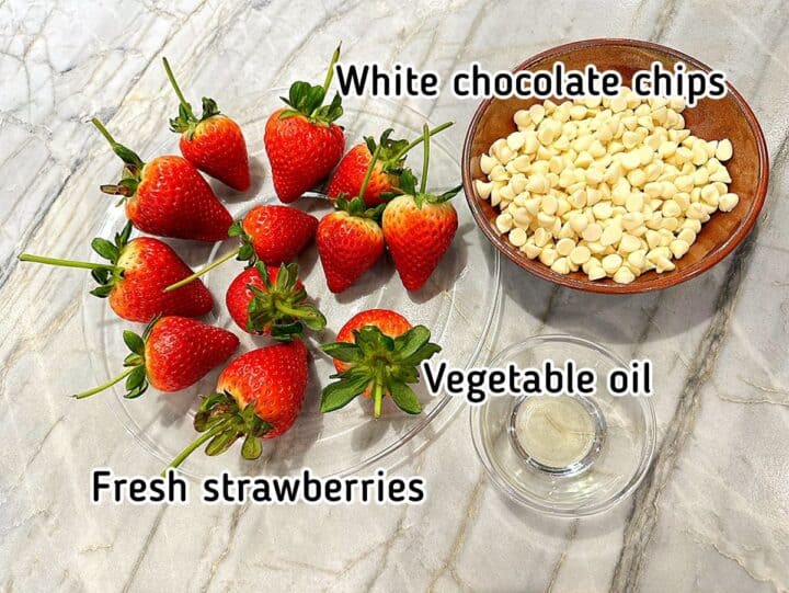Long-stem red strawberries, bowl of white chocolate chips, and small clear bowl of vegetables oil on marble background 