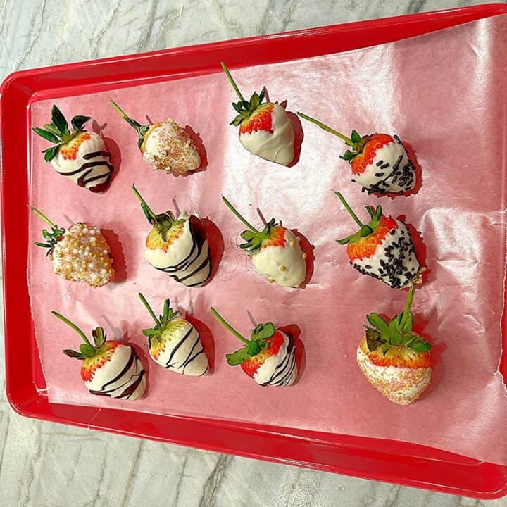 a dozen decorated white-chocolate strawberries on a pink-red sheet pan lined with wax paper.
