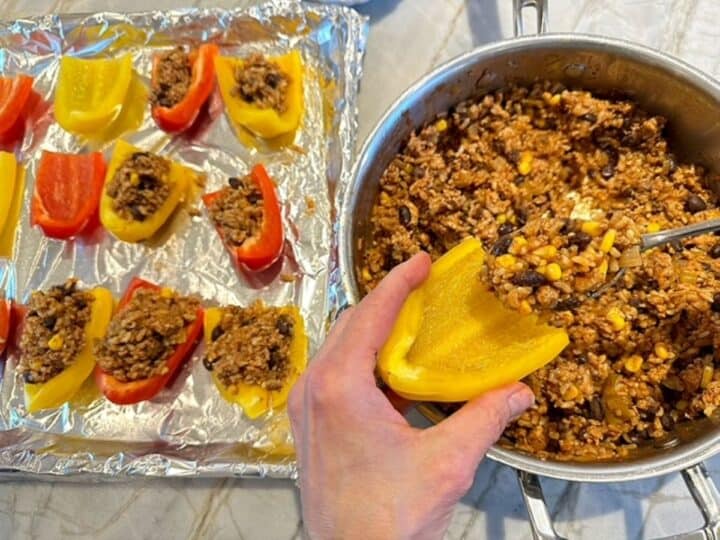 Hand holding yellow bell pepper quarter over skillet while spoon puts ground turkey mixture inside it next to sheet pan of stuffed pepper quarters.
