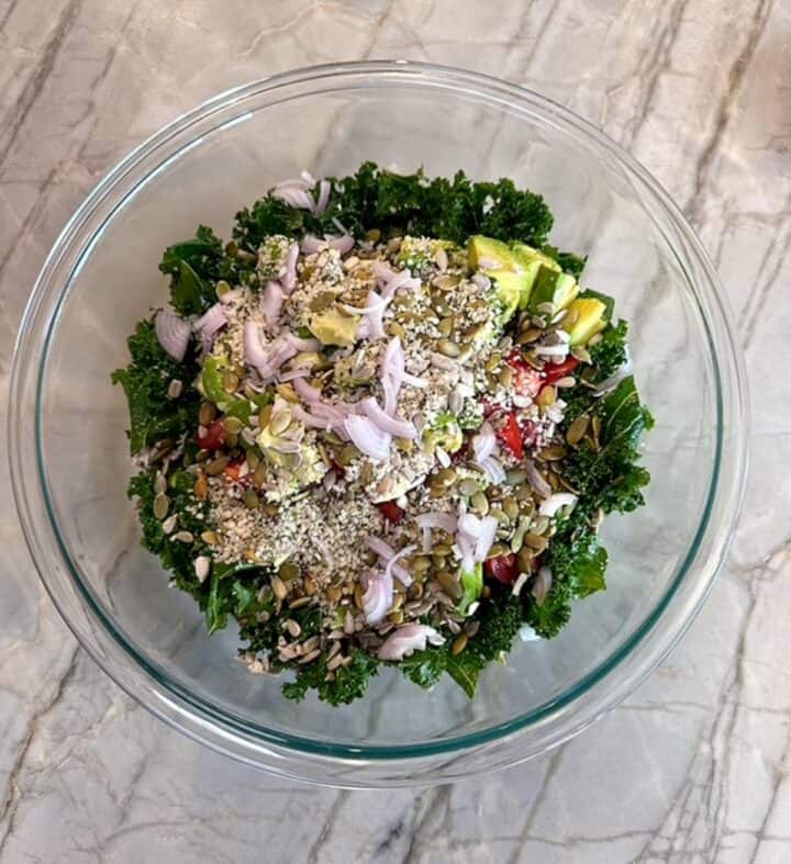 Curly green kale topped with avocado, shallots, seeds, and tomatoes in a clear bowl.