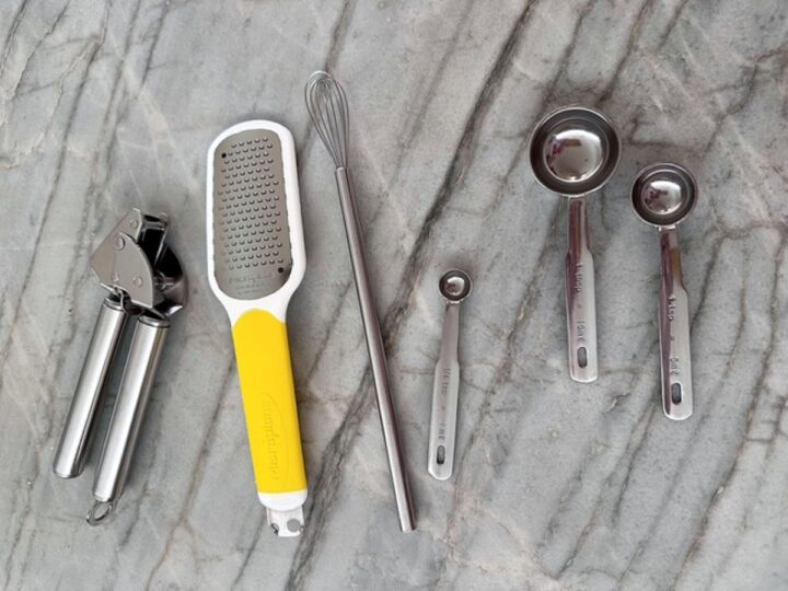 Silver garlic press, long small whisk, yellow handled zester and metal measuring spoons on a gray marble backdrop.