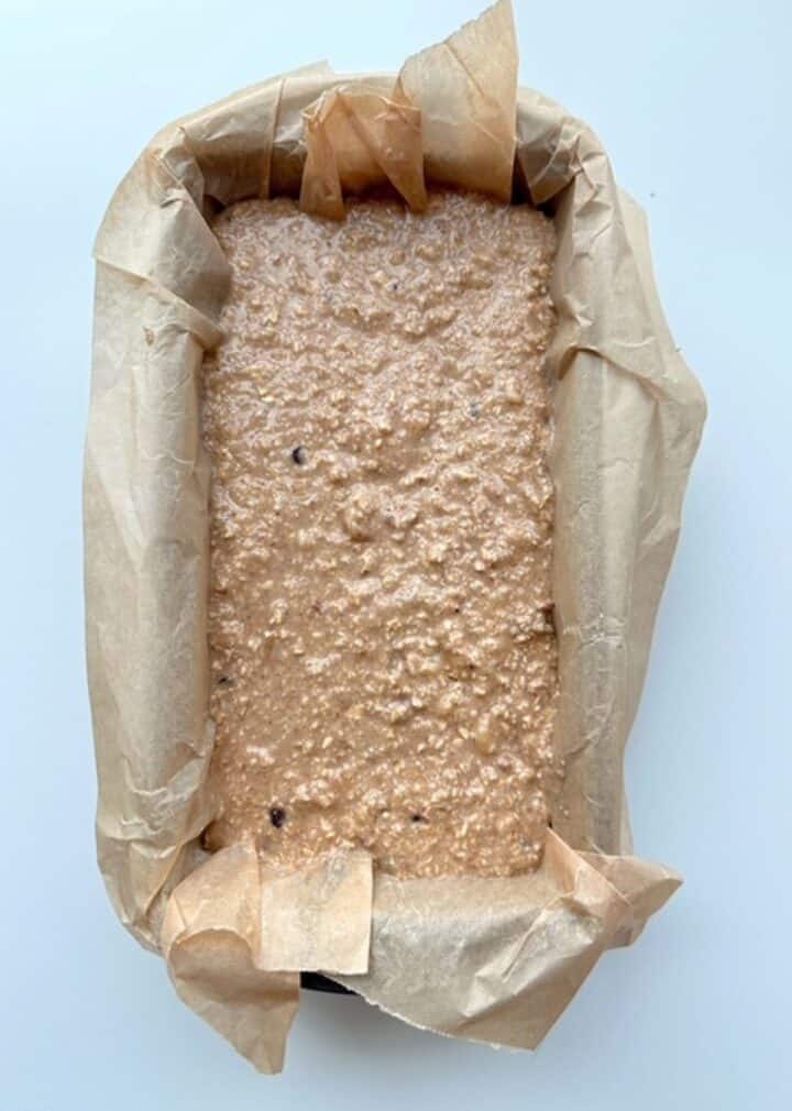 Loaf pan lined in brown parchment full of nubbly beige oat bread batter. 