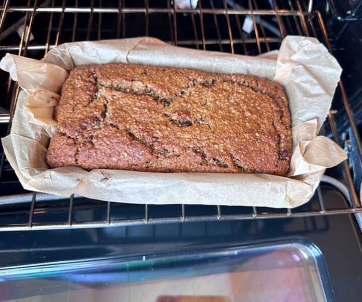 Golden brown loaf of oat flour banana bread shown on an oven rack in a parchment-lined pan.