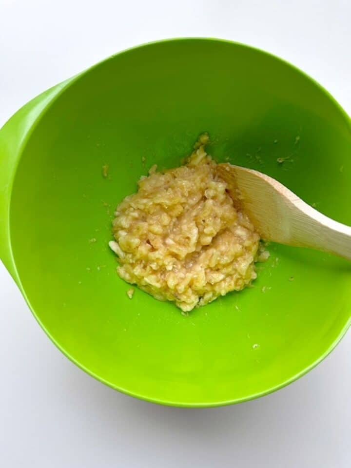 Creamy yellow mashed bananas in the bottom of a lime green mixing bowl with a wooden spatula.
