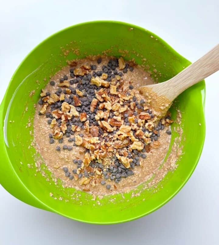 Wooden spatula mixes mini chocolate chips and toasted walnuts into batter in a lime green mixing bowl.