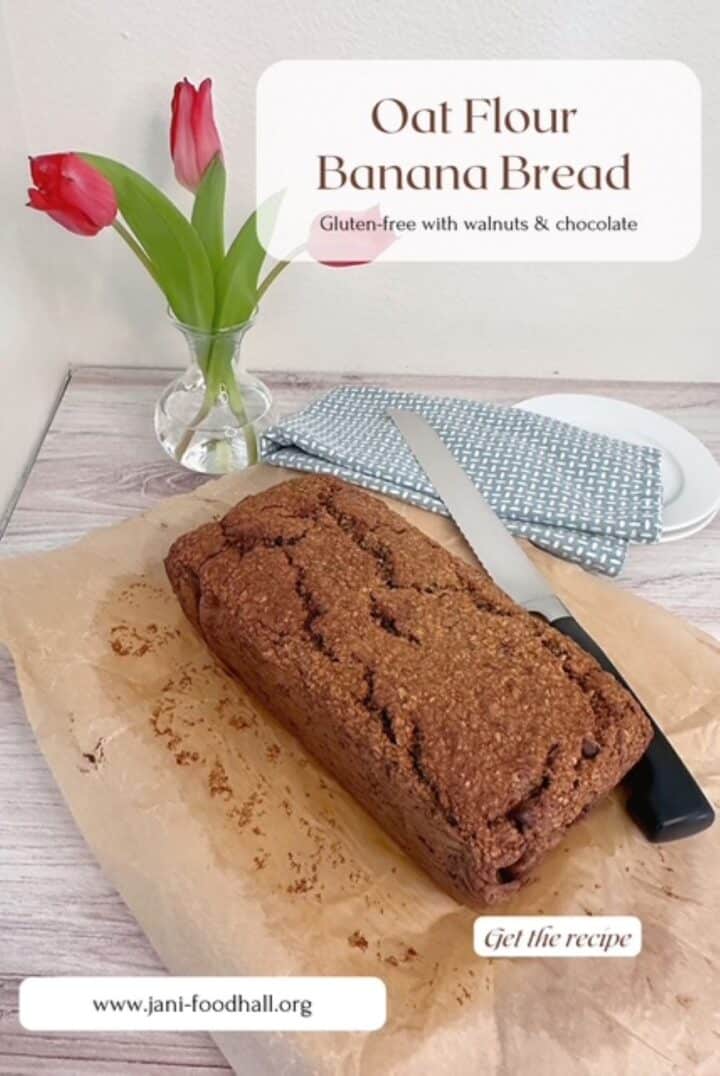 Brown loaf of bread on parchment with a bread knife close by and three dark pink tulips in a vase behind it.