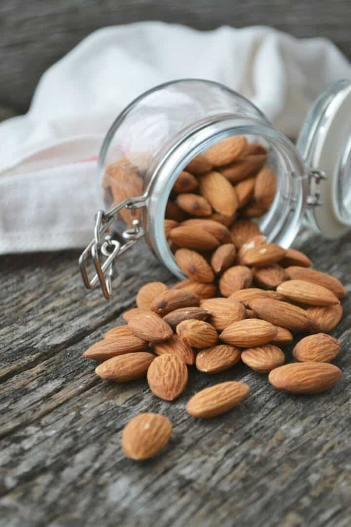 Almonds spilling out of a clear jar onto a wood countertop.