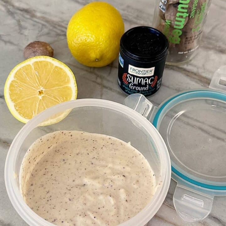 Round opaque storage container holds a creamy tahini sauce with lemons and spice bottles in the background.