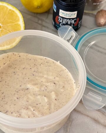 A light tan creamy dip flecked with brown bit is in an opaque container with a yellow cut lemon in the background.