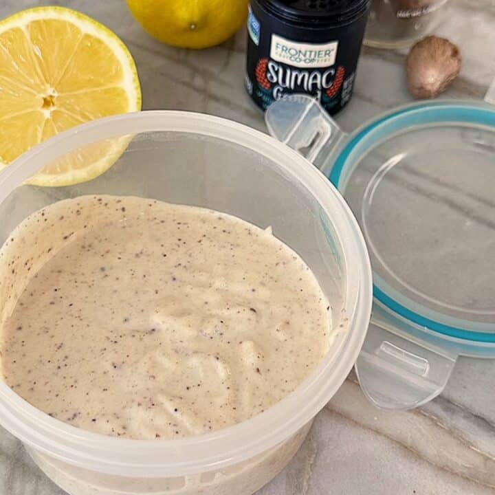A light tan creamy dip flecked with brown bit is in an opaque container with a yellow cut lemon in the background.