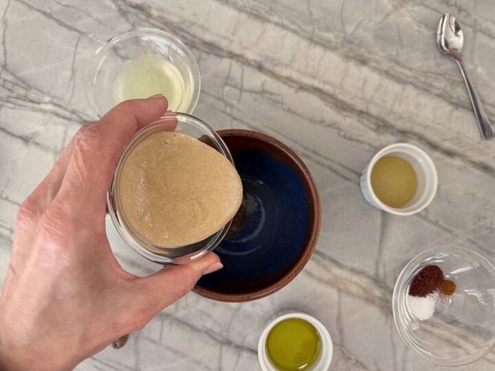 Pouring tan tahini into a brown bowl on a marble counter to make a sauce.