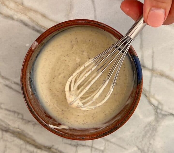 Whisk with creamy bits clinging to it hovers over brown bowl filled with creamy, thick tahini sauce.
