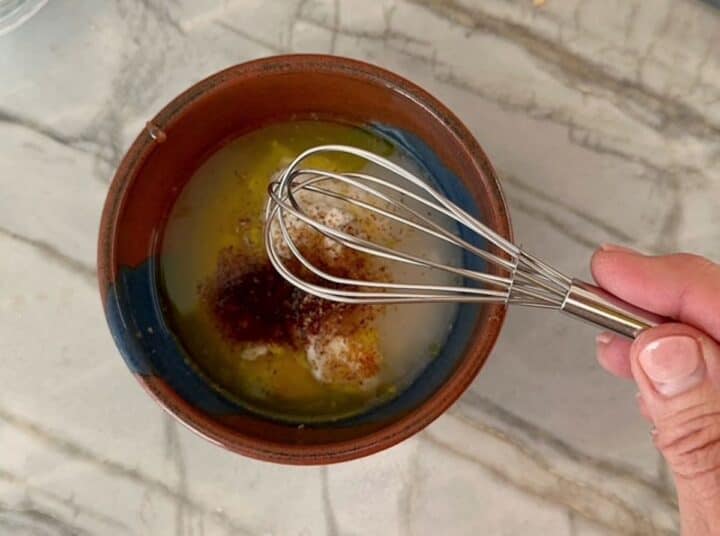 Metal whisk hovers over brown bowl full of various liquid ingredients.