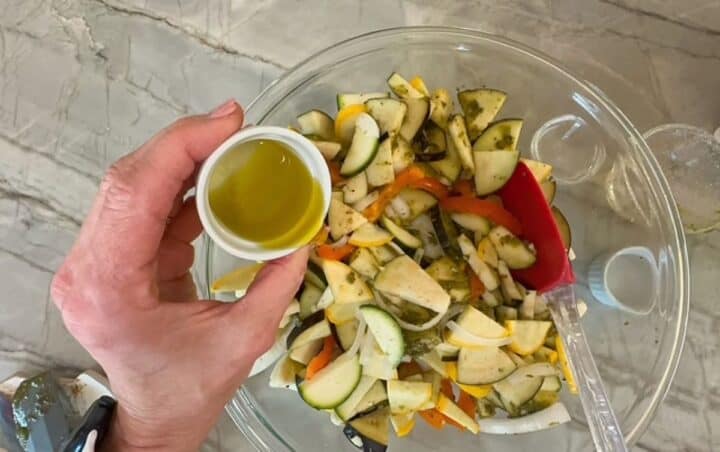 Hand holding a small white ramekin full of green olive oil to pour over cut veggies in a clear bowl.