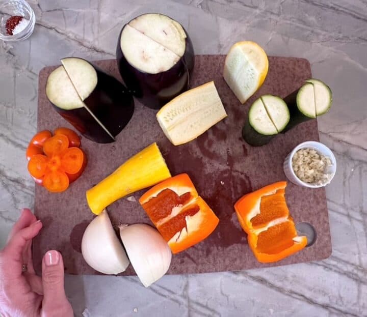 Several tender veggies including eggplant, yellow squash, and orange bell pepper are partially prepped for roasting on a brown cutting board.
