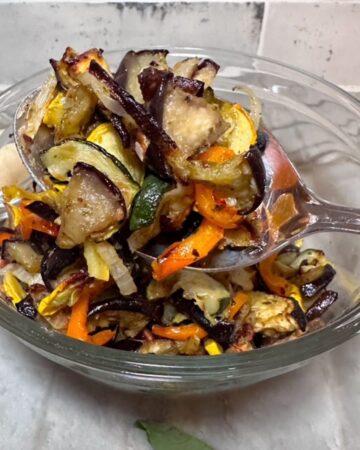 Close view of a clear bowl full of colorful pesto roasted vegetables with a large spoonful front and center.