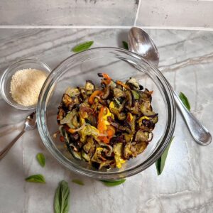 Clear bowl with softened, browning roasted veggies with grated Parmesan and a big spoon for serving.