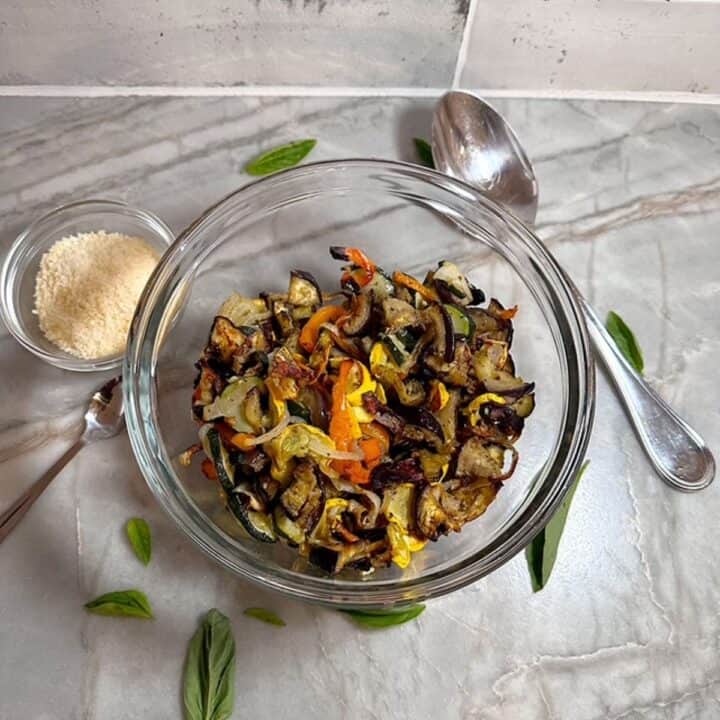 Clear bowl with softened, browning roasted veggies with grated Parmesan and a big spoon for serving.