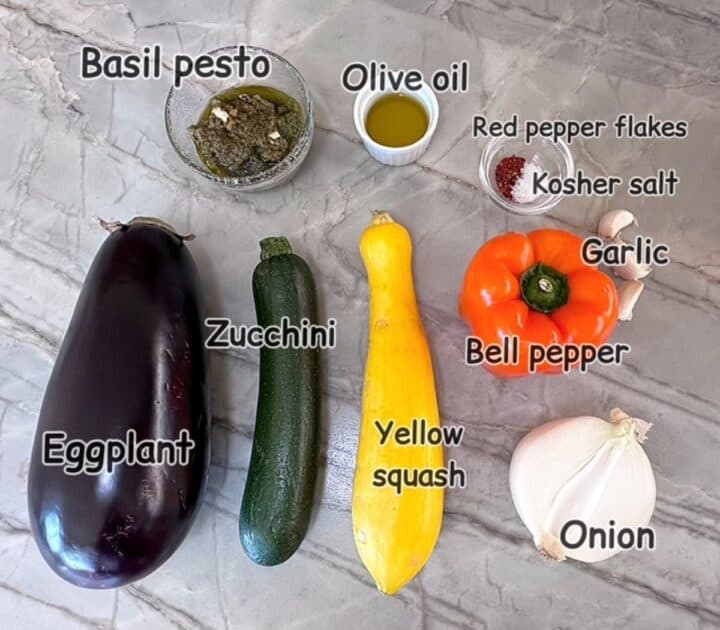 Purple eggplant, green zucchini, yellow squash, orange bell pepper plus pesto and seasoning on a marble countertop.
