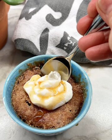 A metal spoon dips into amber Honey Almond Butter Cake topped with white yogurt dollop in a green ramekin.