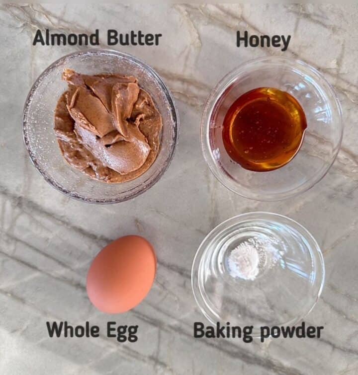 Almond butter, honey, whole brown egg, and white baking powder labeled on a gray marble-swirled countertop.