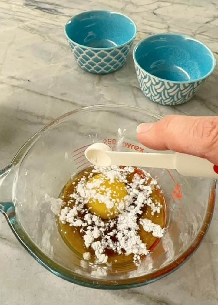 Small white measuring spoon depositing white baking powder over a raw egg in a clear measuring cup.