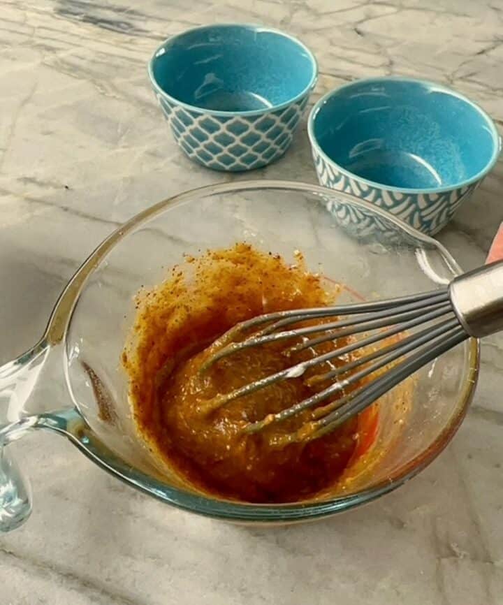 Gray silicone whisk covered in sticky brown honey almond butter batter in a clear measuring cup.