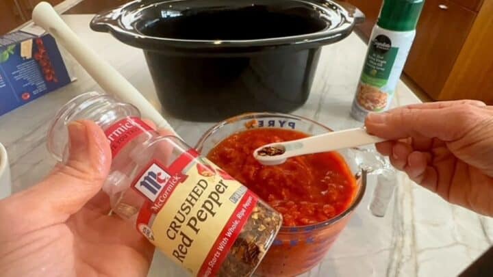 Jar of red pepper flakes held in front of a bowl of red tomato sauce with white measuring spoon.